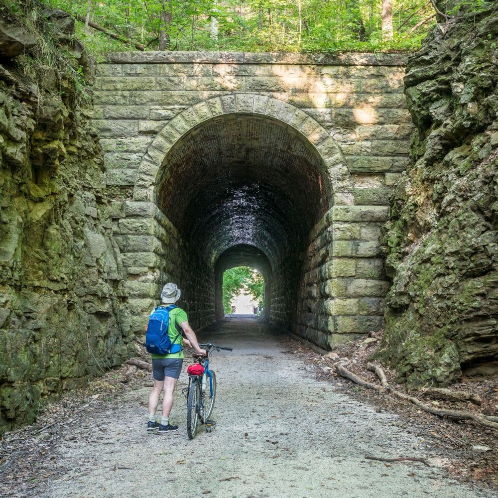 A cyclist stands at the entrance of a stone tunnel surrounded by lush greenery.