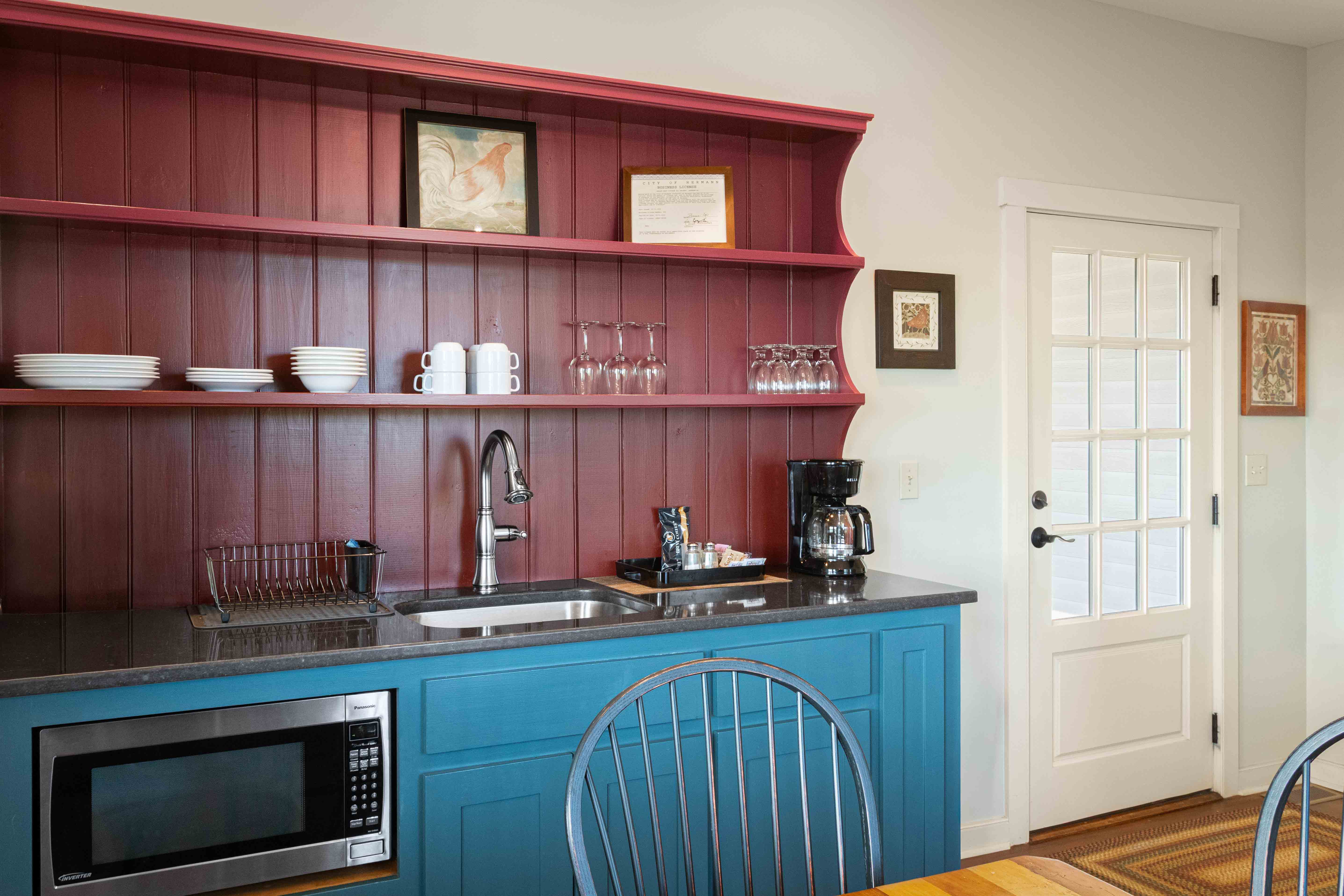 A modern kitchen corner featuring a blue lower cabinet, red shelving with white dishware, and a door leading outside.