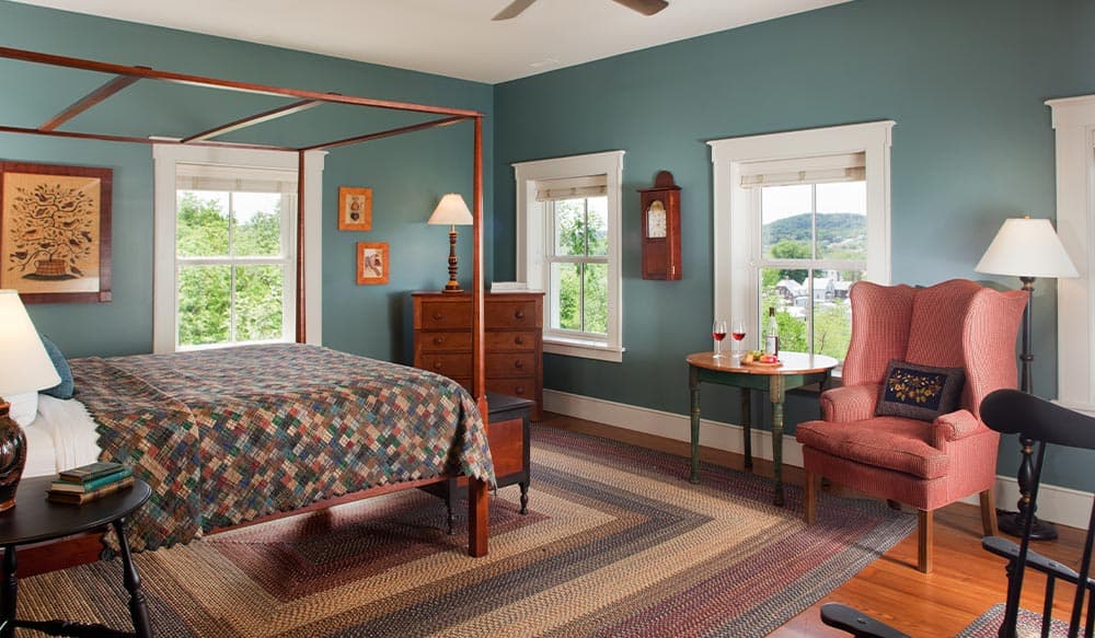 A cozy bedroom featuring a four-poster bed, colorful quilt, armchair, and windows with a scenic view.