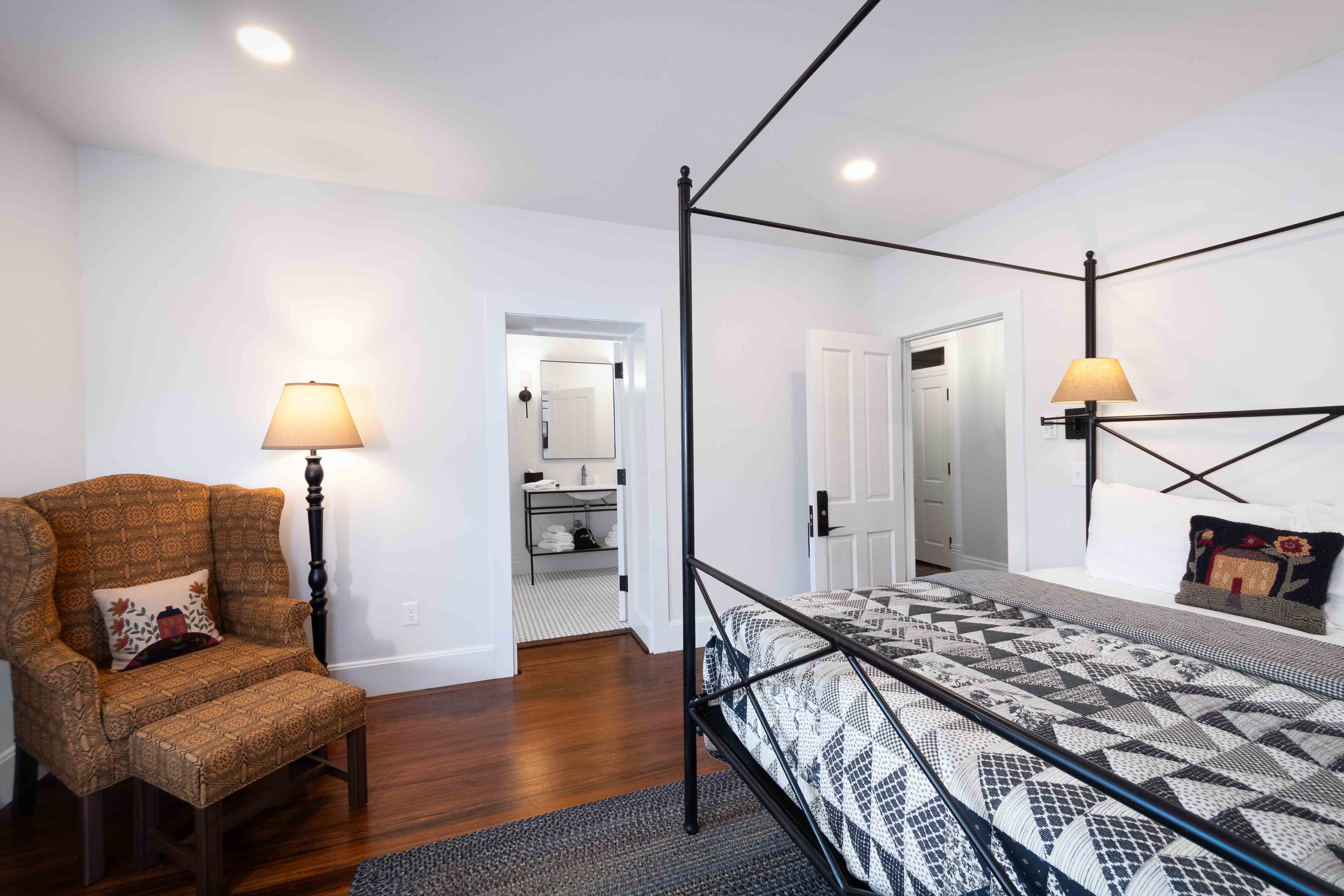 A modern bedroom featuring a four-poster bed, patterned quilt, armchair, and an ensuite bathroom door.