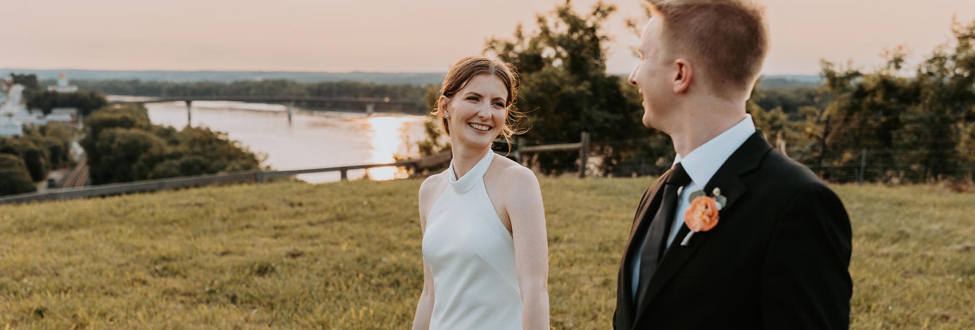 A bride and groom smile at each other, standing on a grassy hill with a river and sunset in the background.