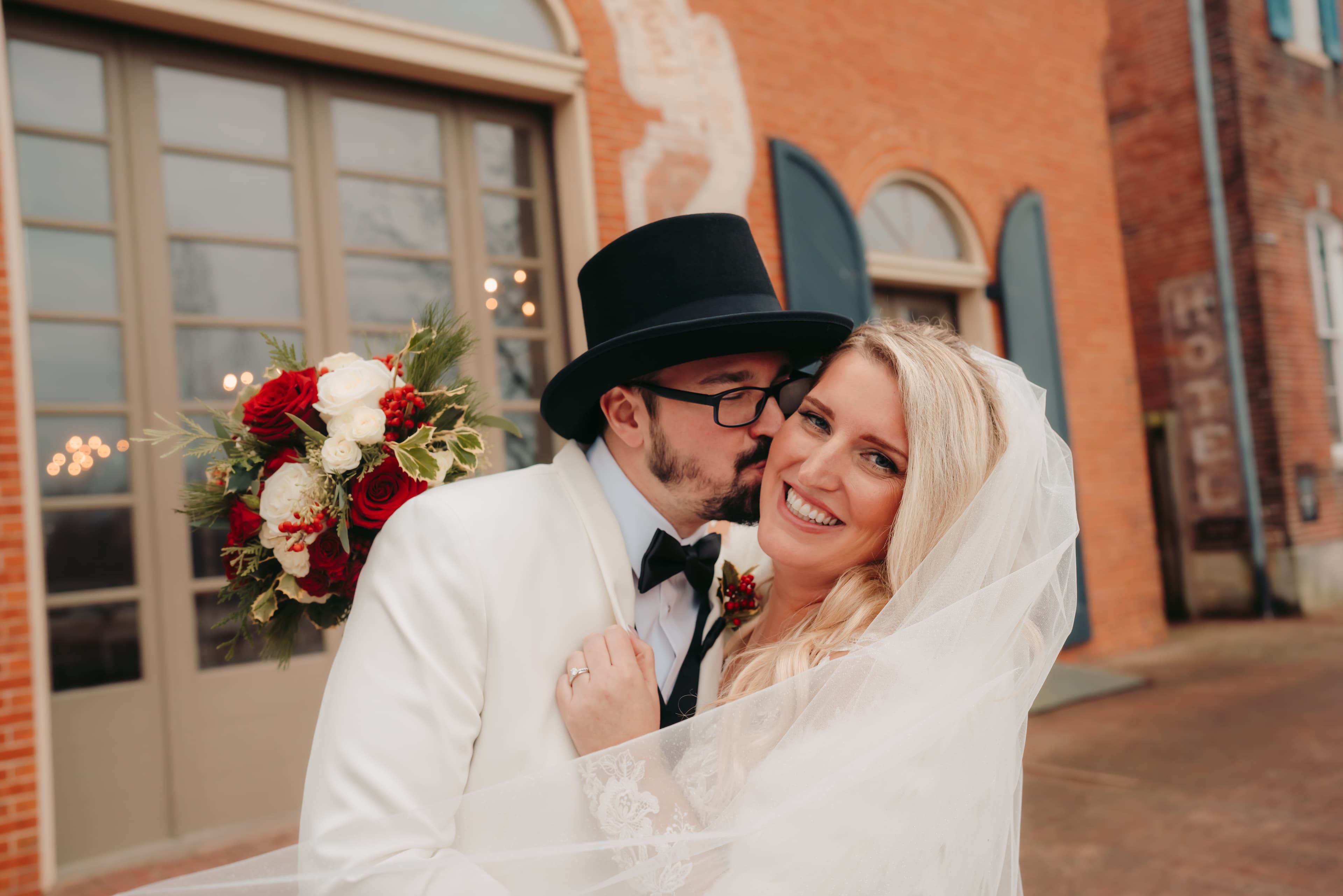 A bride and groom share a joyful moment outside a historic building, with the bride holding a bouquet of red and white flowers.