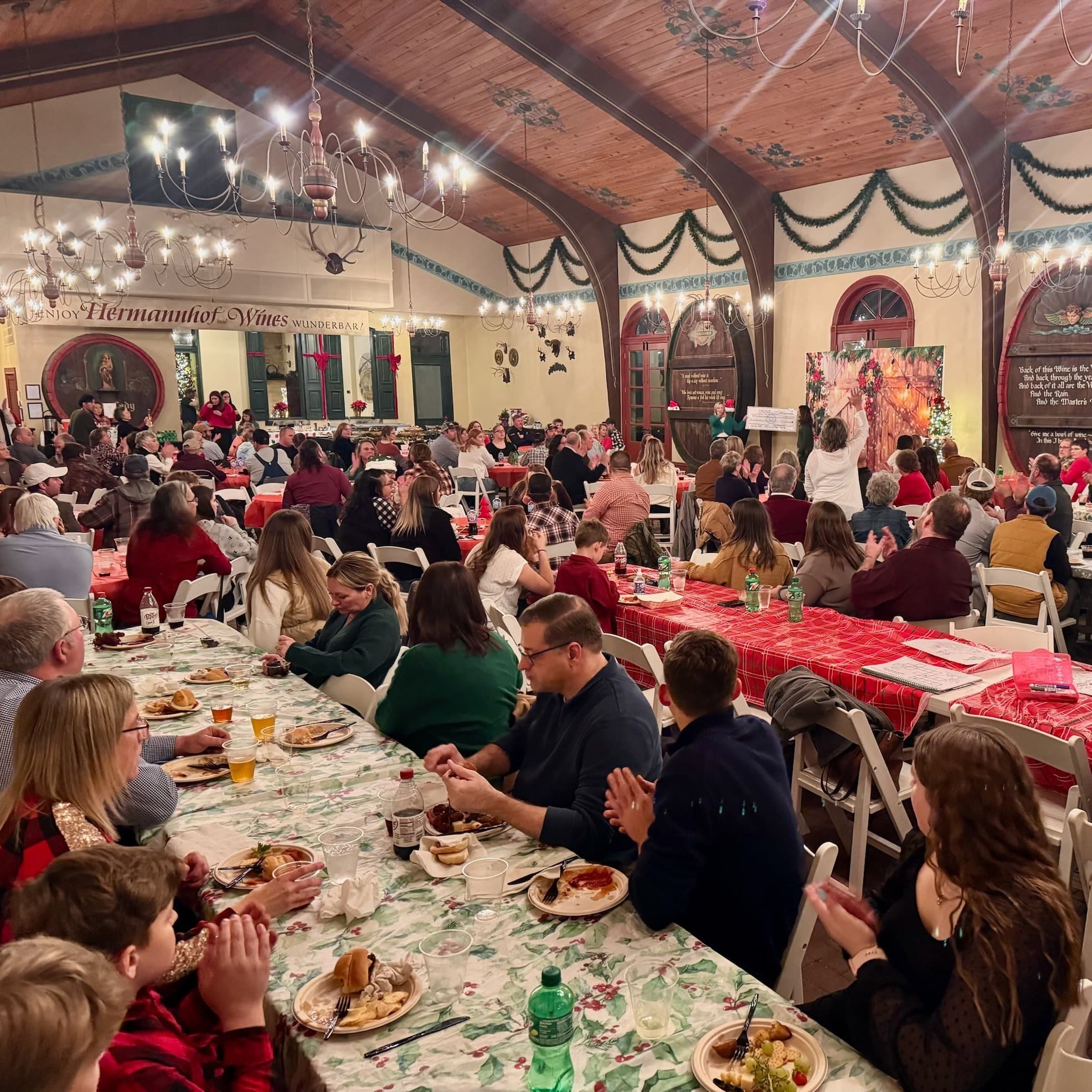 A festive gathering with people seated at long tables, enjoying a meal and celebrating in a decorated hall.