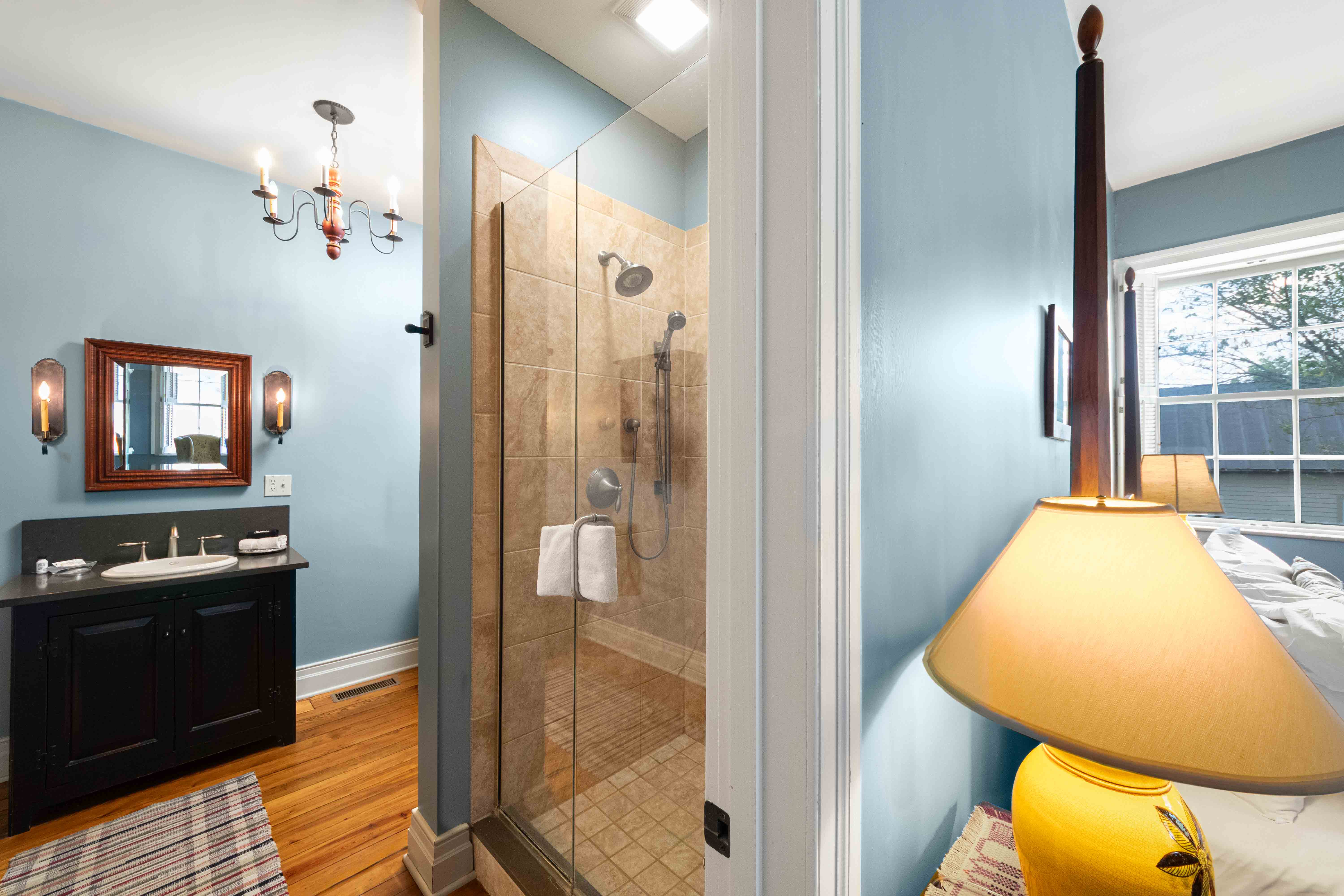 A well-lit bathroom featuring a glass shower, dark cabinetry, and a cozy bedroom area with a lamp.