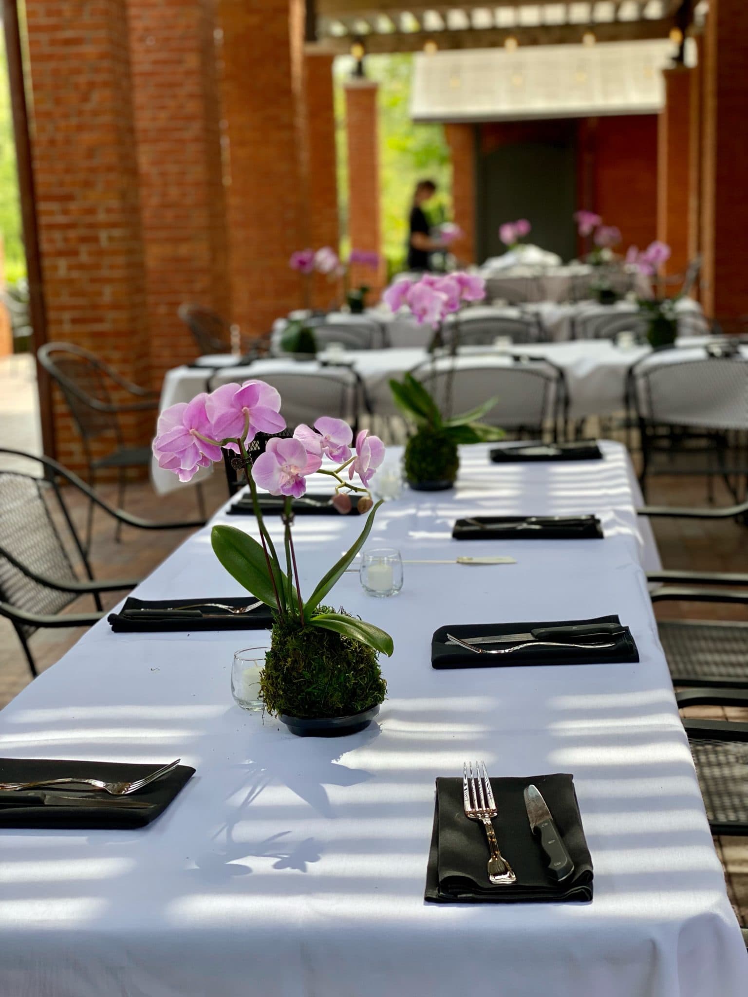 A beautifully set dining table with pink orchids and black napkins in a sunlit outdoor setting.