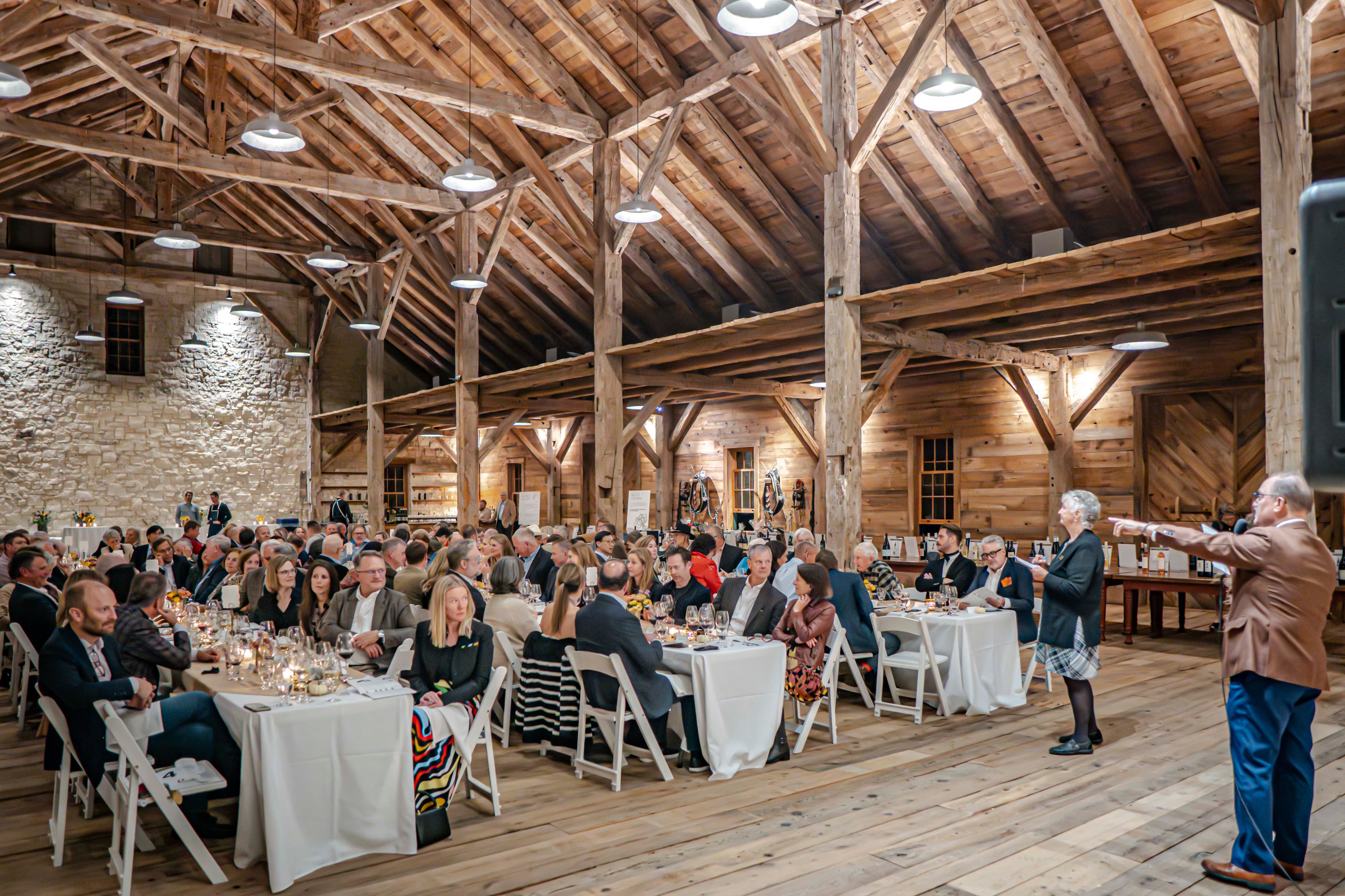 A large group of attendees sits at tables in a rustic venue during a formal event.