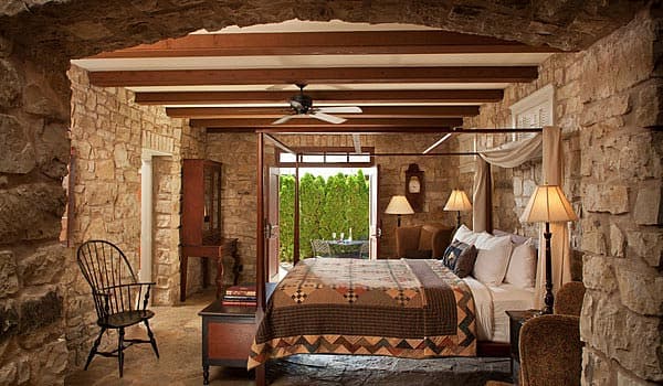 A cozy bedroom featuring a stone wall, wood beams, a four-poster bed, and a view of greenery through glass doors.