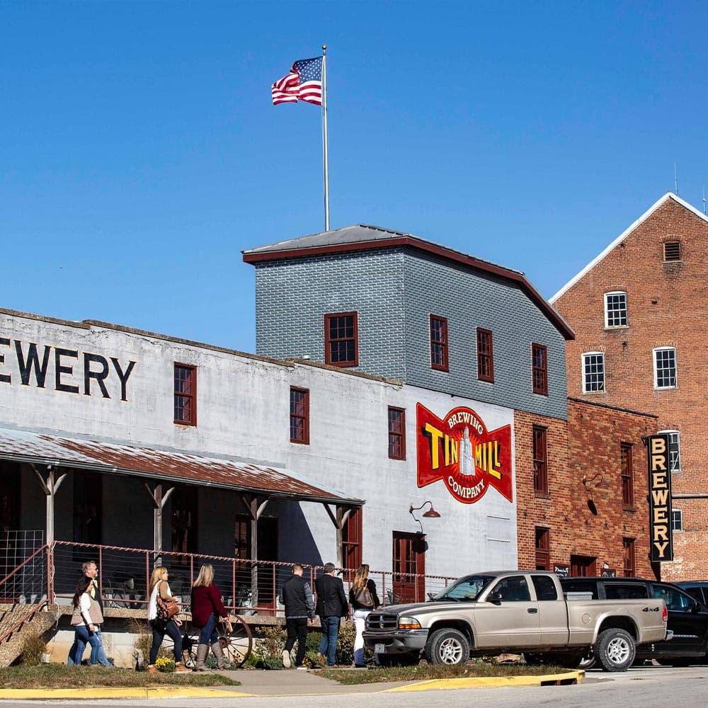 A group of people walks towards the Tin Mill Brewery, featuring an American flag and historic buildings.