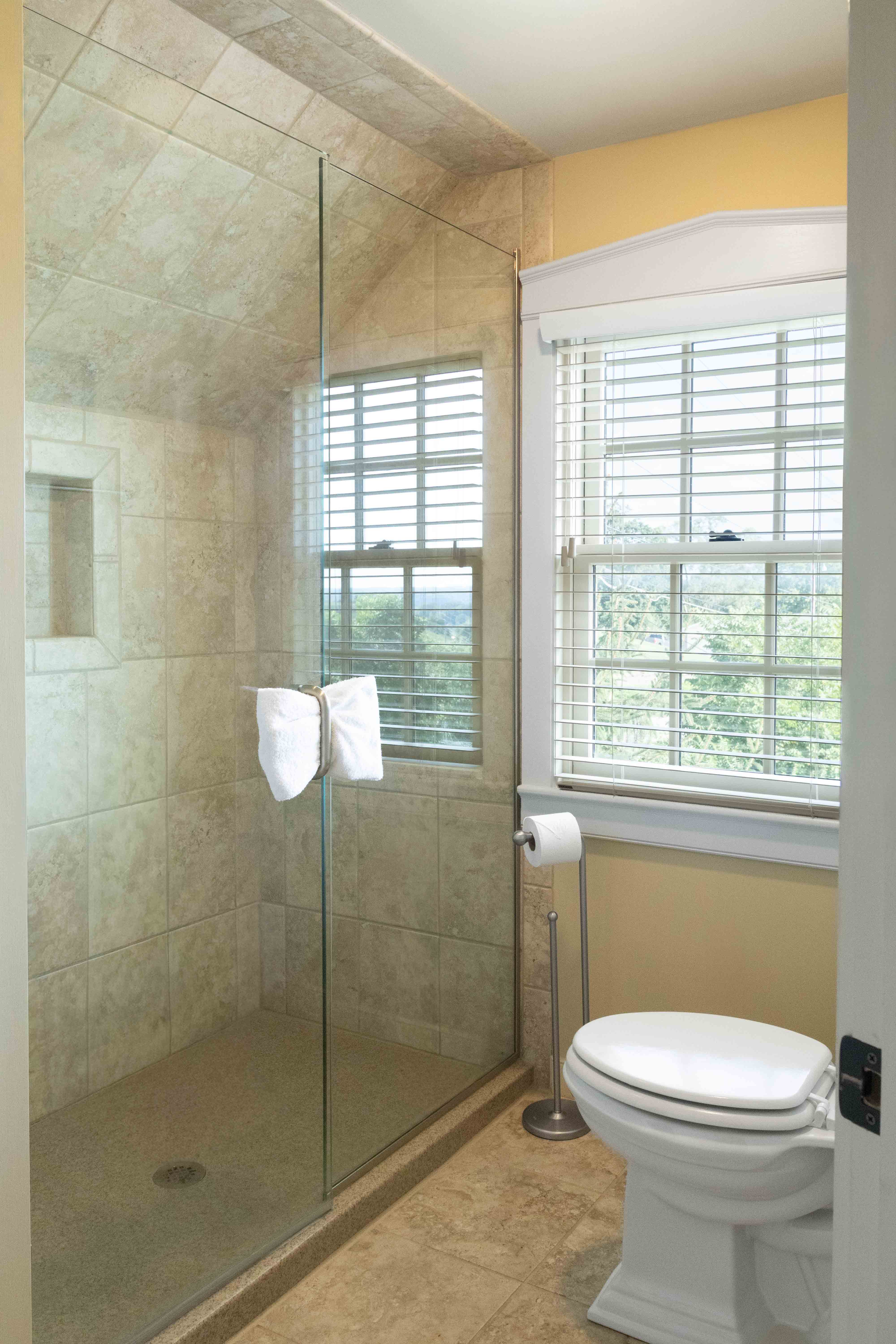 A modern bathroom featuring a glass shower, toilet, and window with blinds.