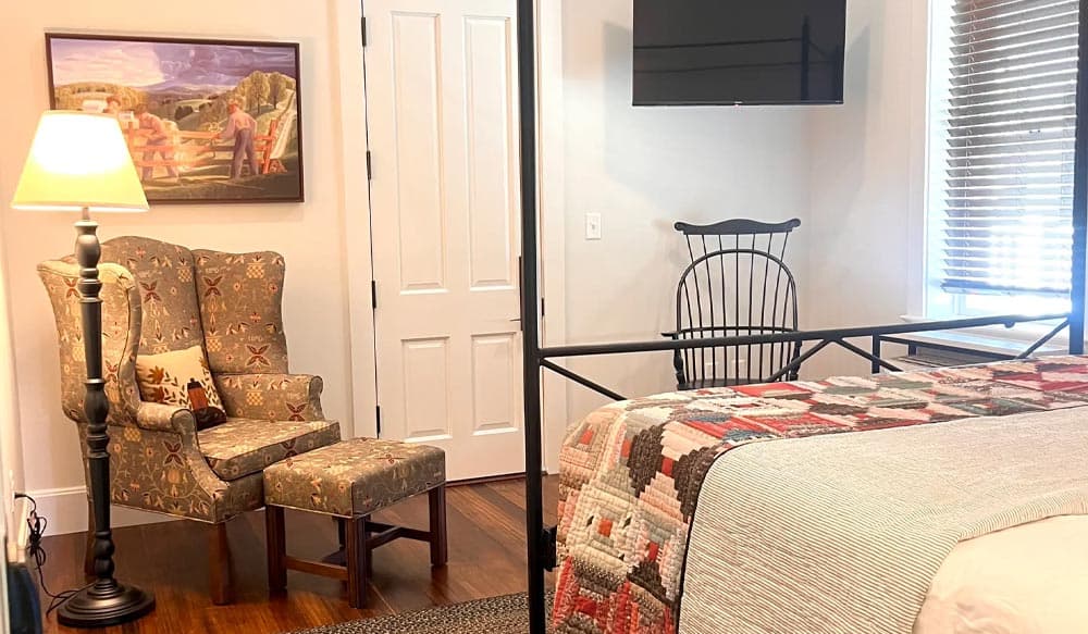 A cozy bedroom featuring a patterned quilt, an armchair with an ottoman, a wall painting, and a wall-mounted TV.