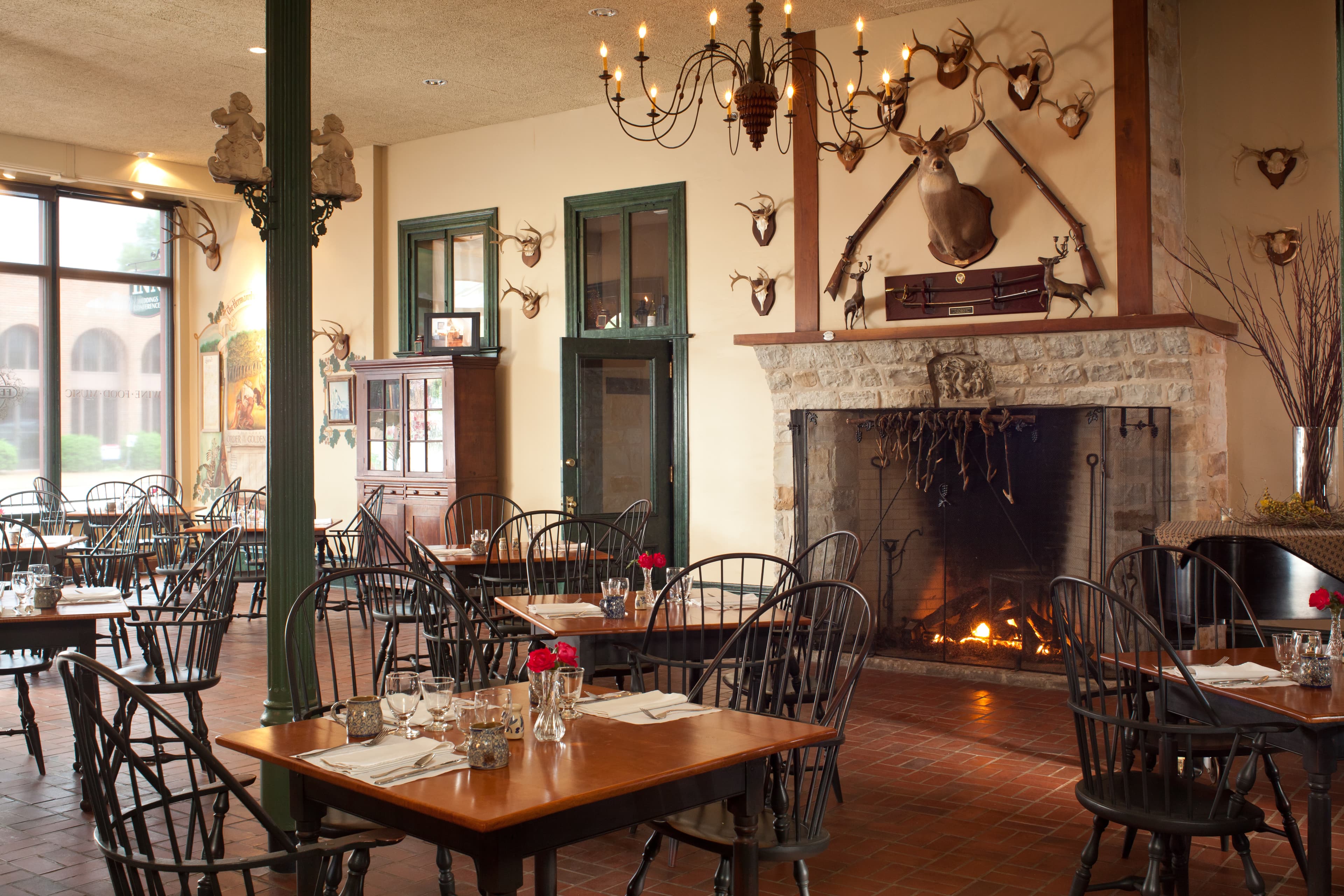 A cozy restaurant interior featuring wooden tables, black chairs, a fireplace, and rustic decor with mounted trophies.