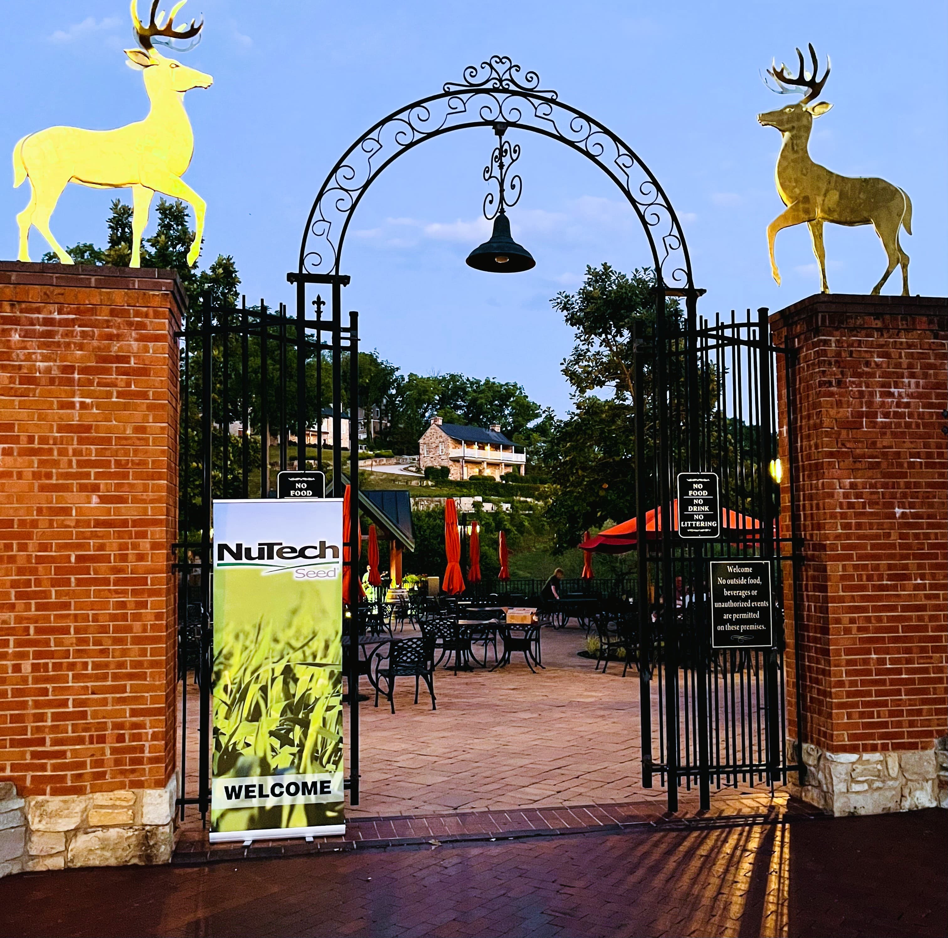 A decorative gate with yellow deer statues and a "Welcome" banner leads into a dining area surrounded by greenery.