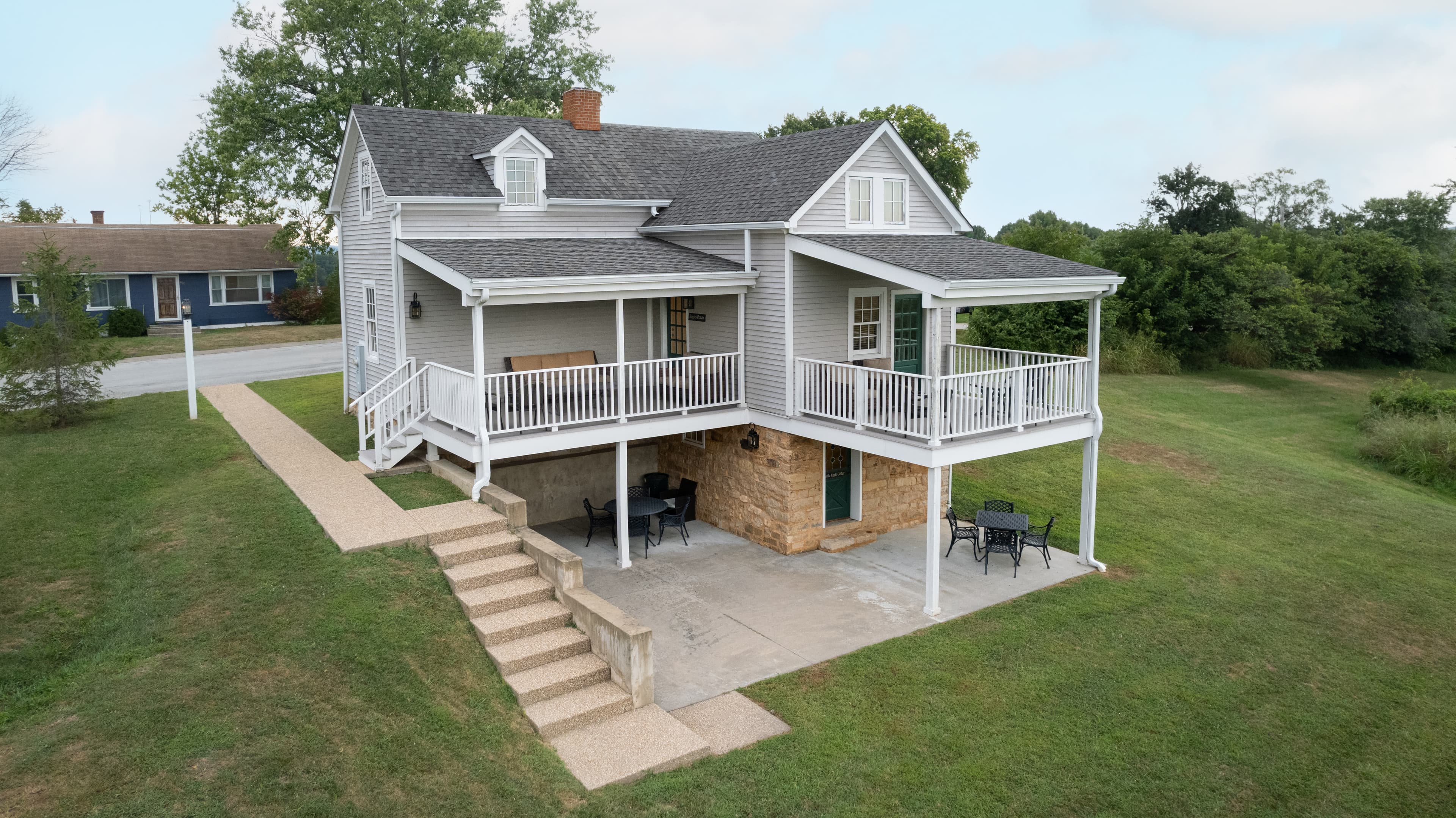A two-story house with a wraparound porch and stone foundation, surrounded by grassy areas.