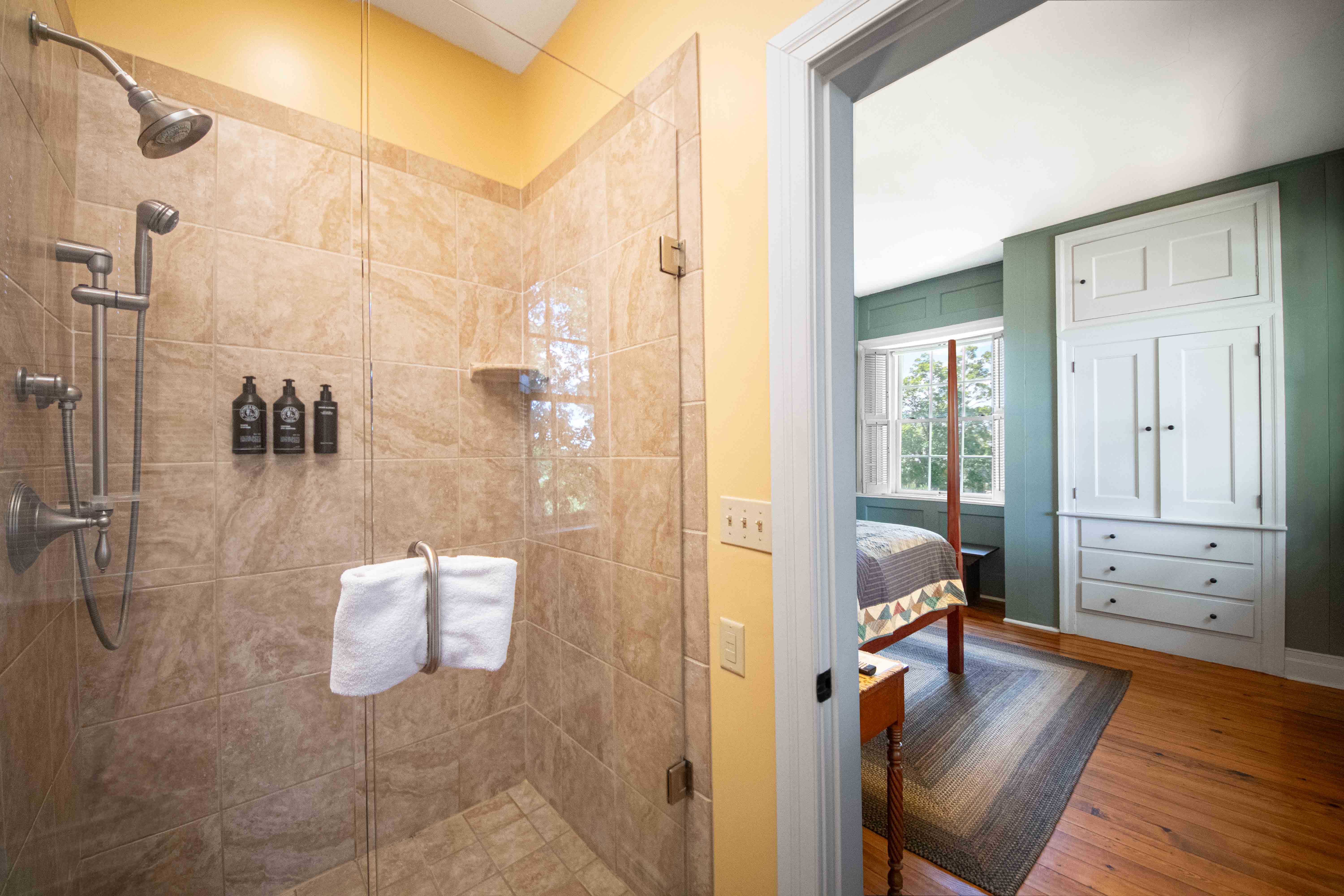 A modern bathroom featuring a glass shower enclosure and an adjoining bedroom with a green wall.