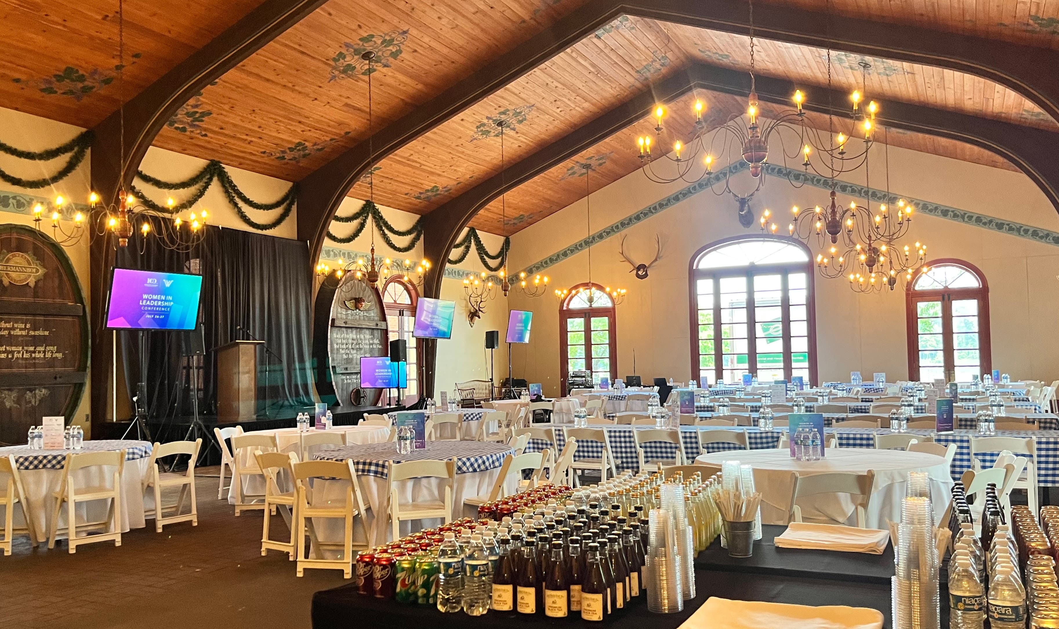 A spacious event hall set up with white tables, chairs, and a stage for presentations, featuring decorative chandeliers and a variety of beverages displayed on a table.