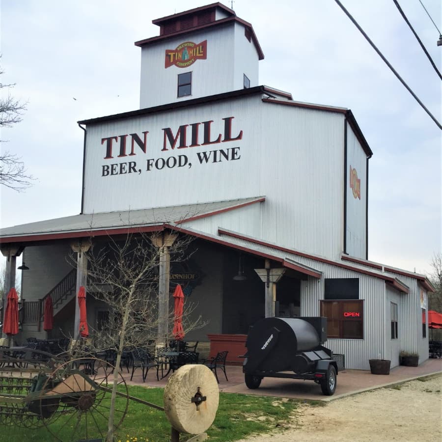 Exterior of the Tin Mill building, featuring signage that reads "TIN MILL BEER, FOOD, WINE" and outdoor seating with umbrellas.
