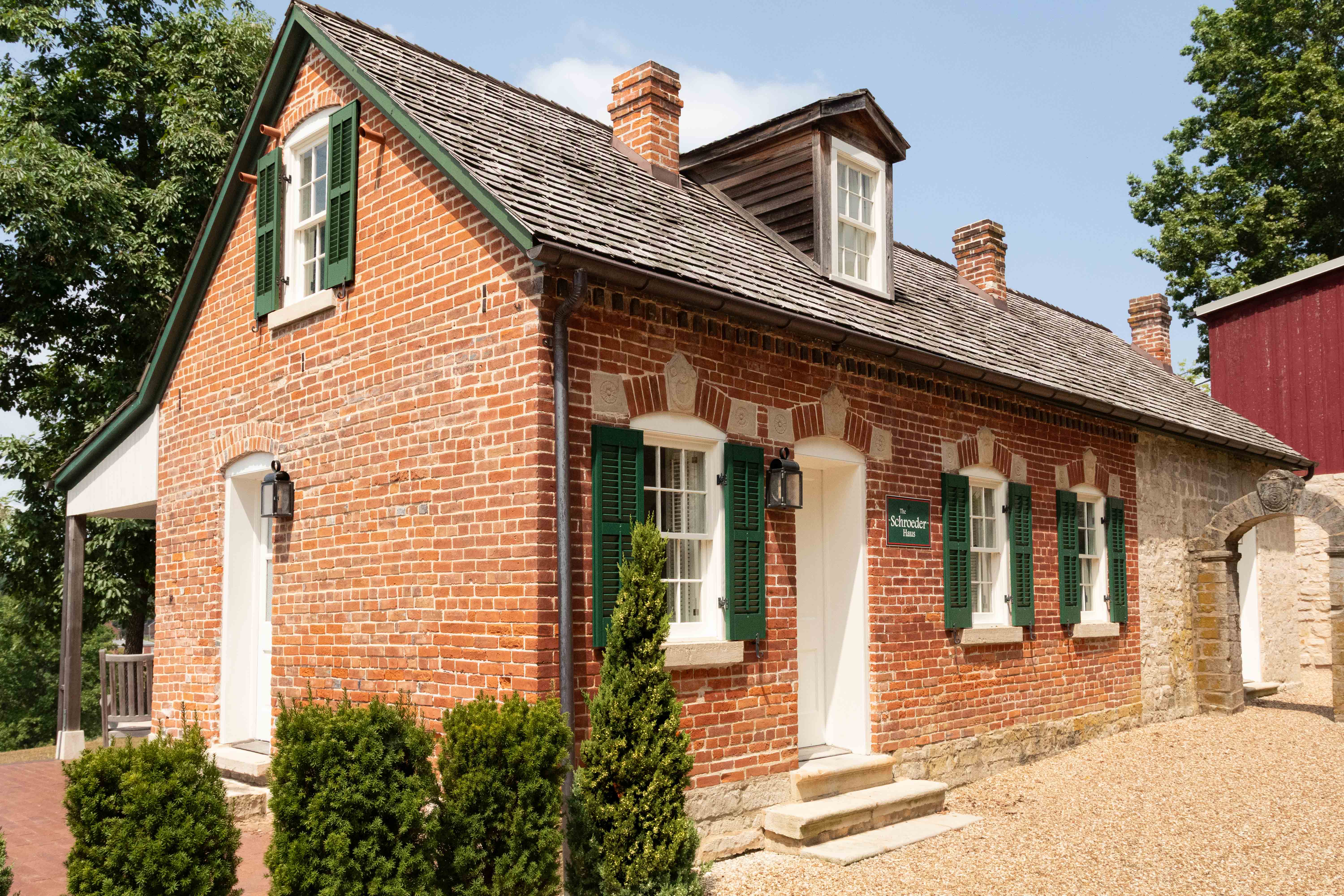A brick house with green shutters and a shingled roof surrounded by greenery.