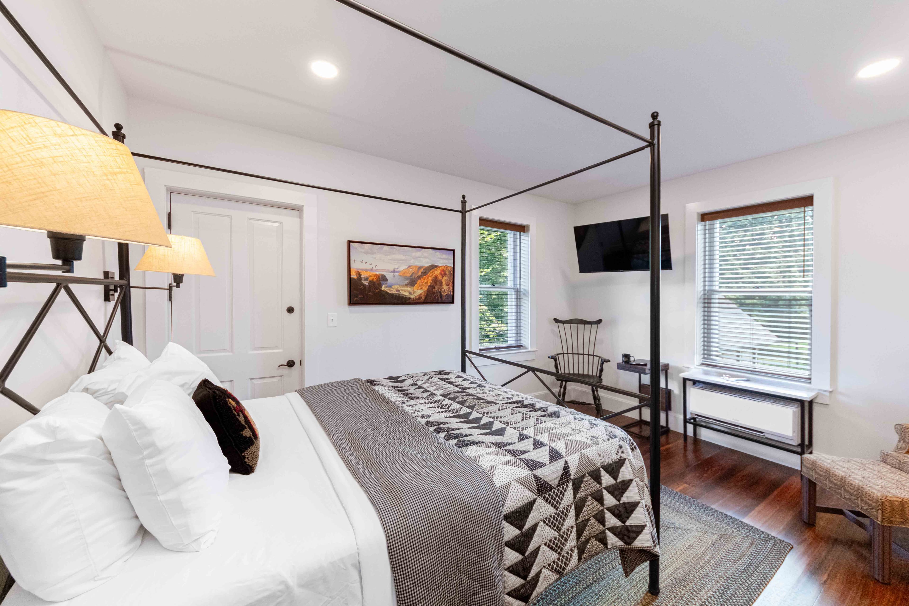 A cozy bedroom featuring a four-poster bed, patterned quilt, a TV, and natural light from two windows.