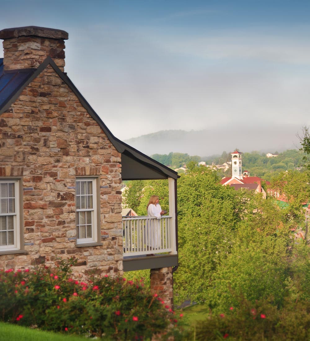 A person in a white robe stands on the balcony of a stone building overlooking a scenic view.
