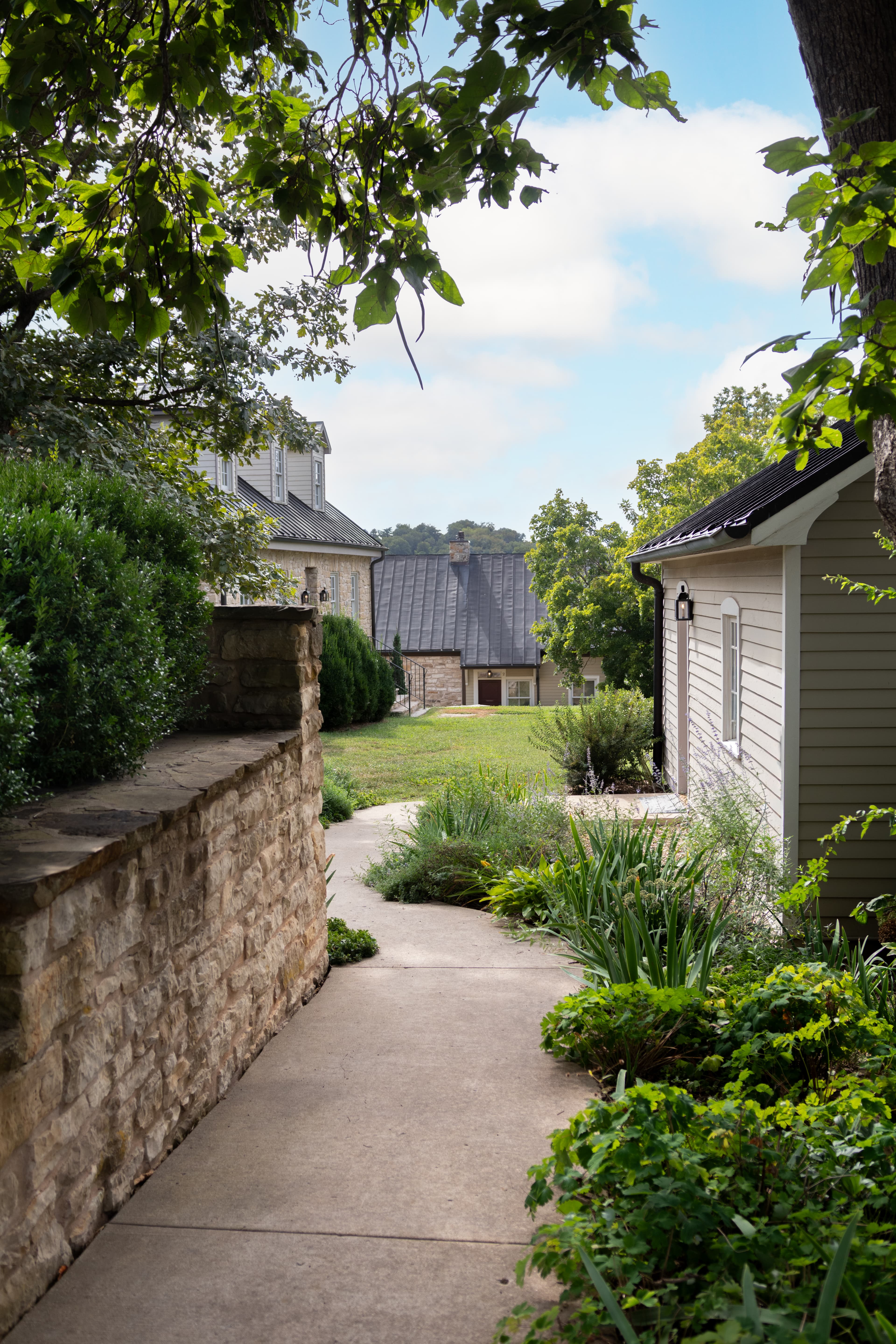 A winding pathway leads through lush greenery toward two houses under a partly cloudy sky.