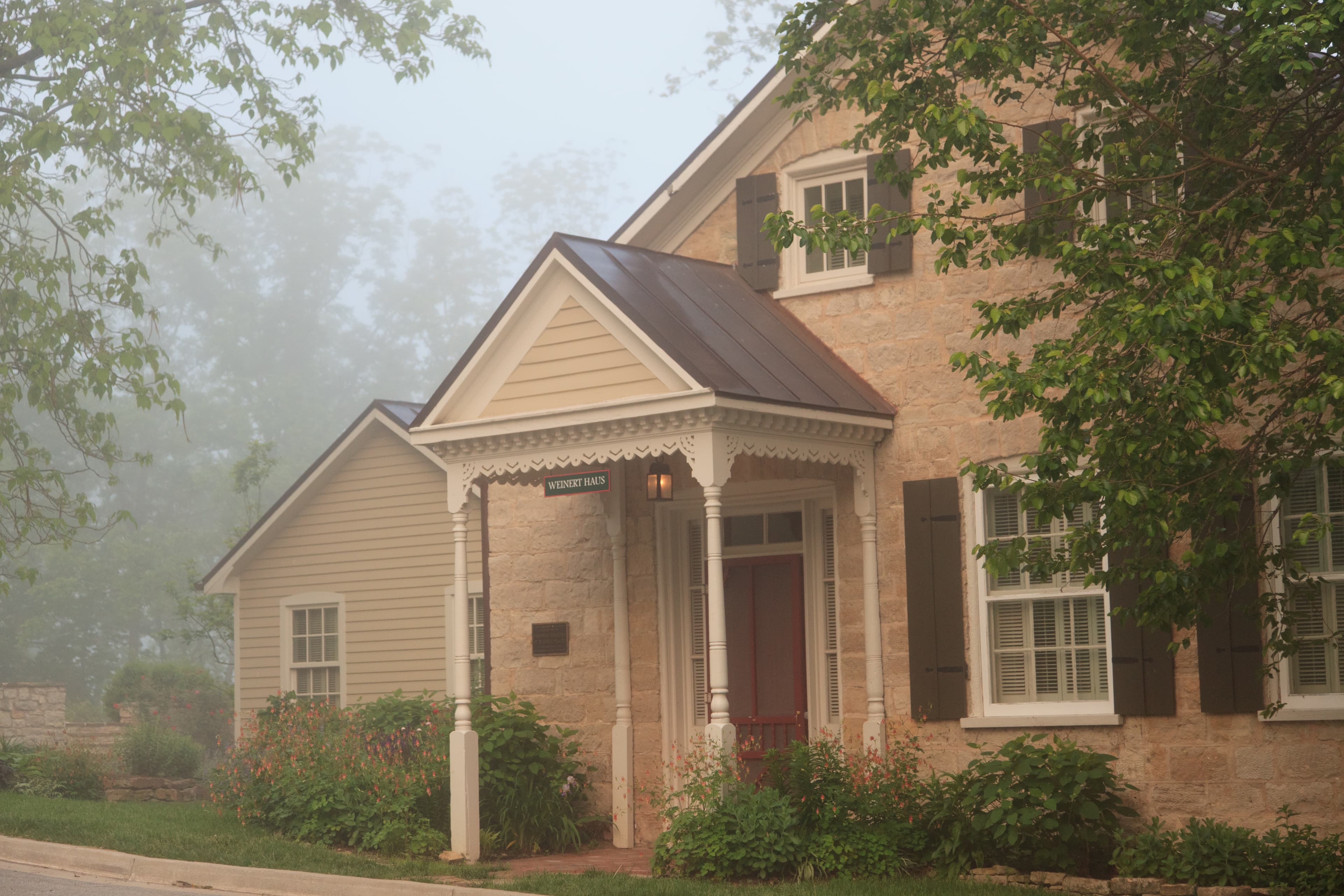 A quaint, two-story stone house partially obscured by fog, featuring a front porch and well-maintained garden.