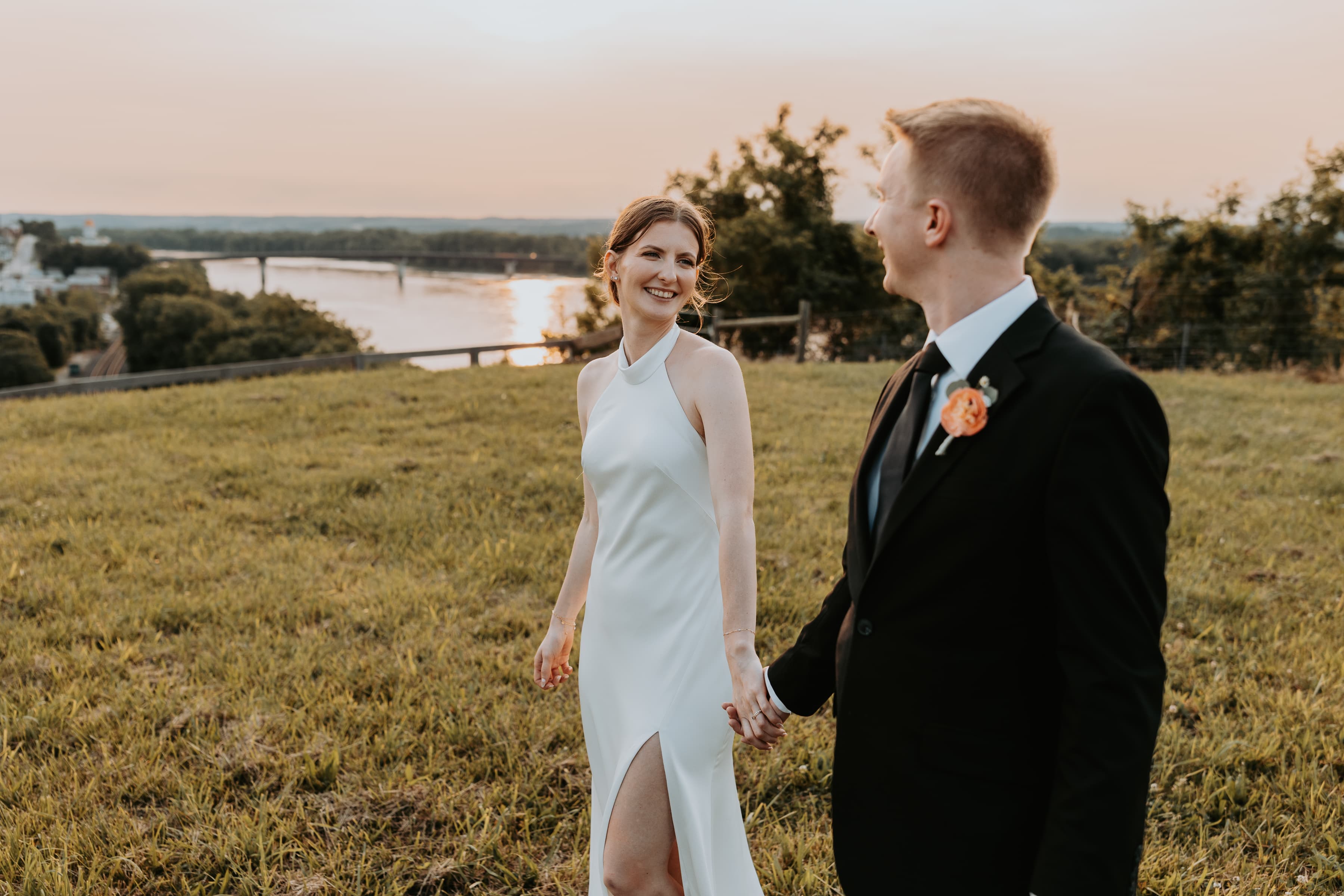 A bride and groom hold hands while enjoying a sunset view by the river.