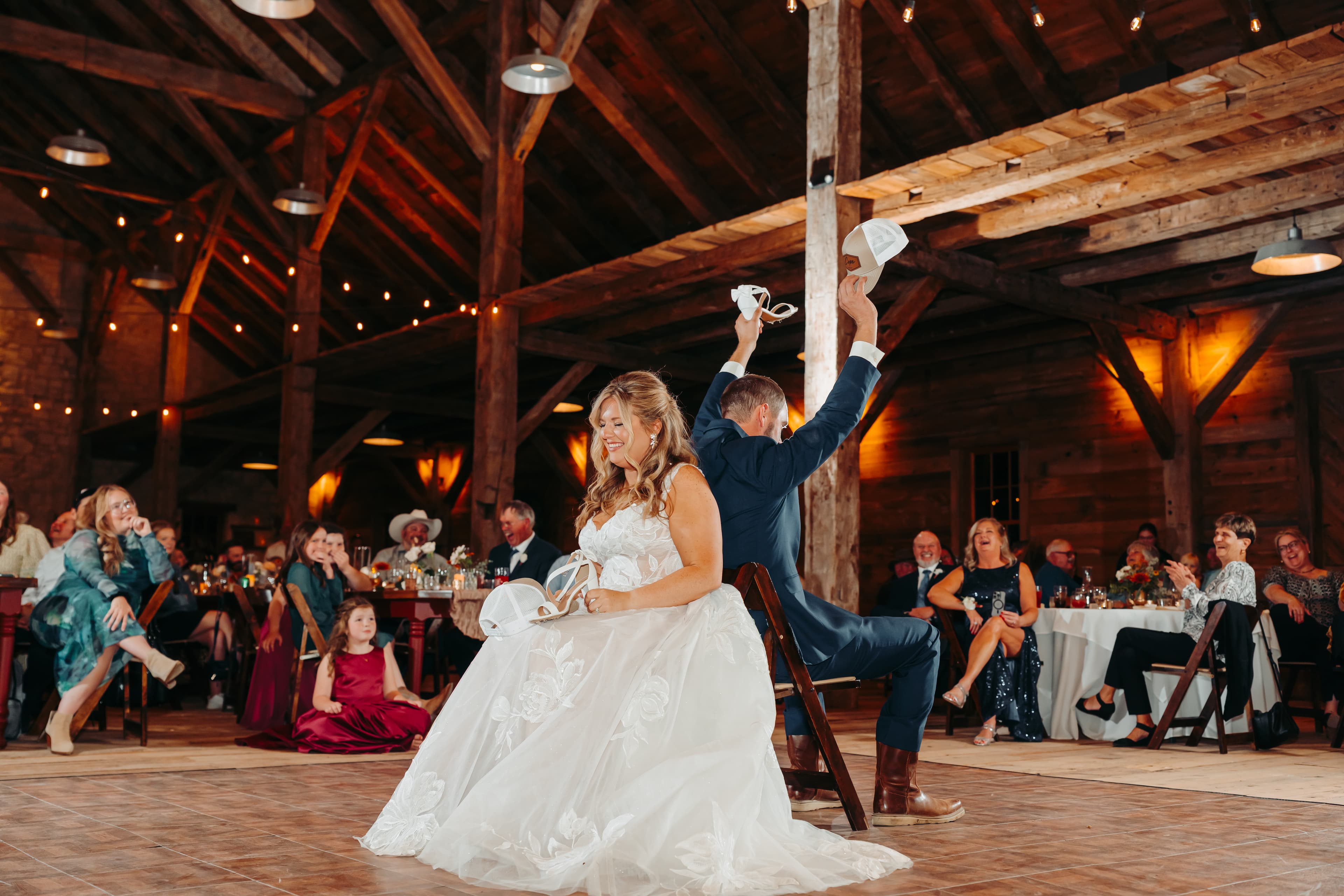 A bride and groom are seated back-to-back, playfully holding up shoes during their wedding reception.
