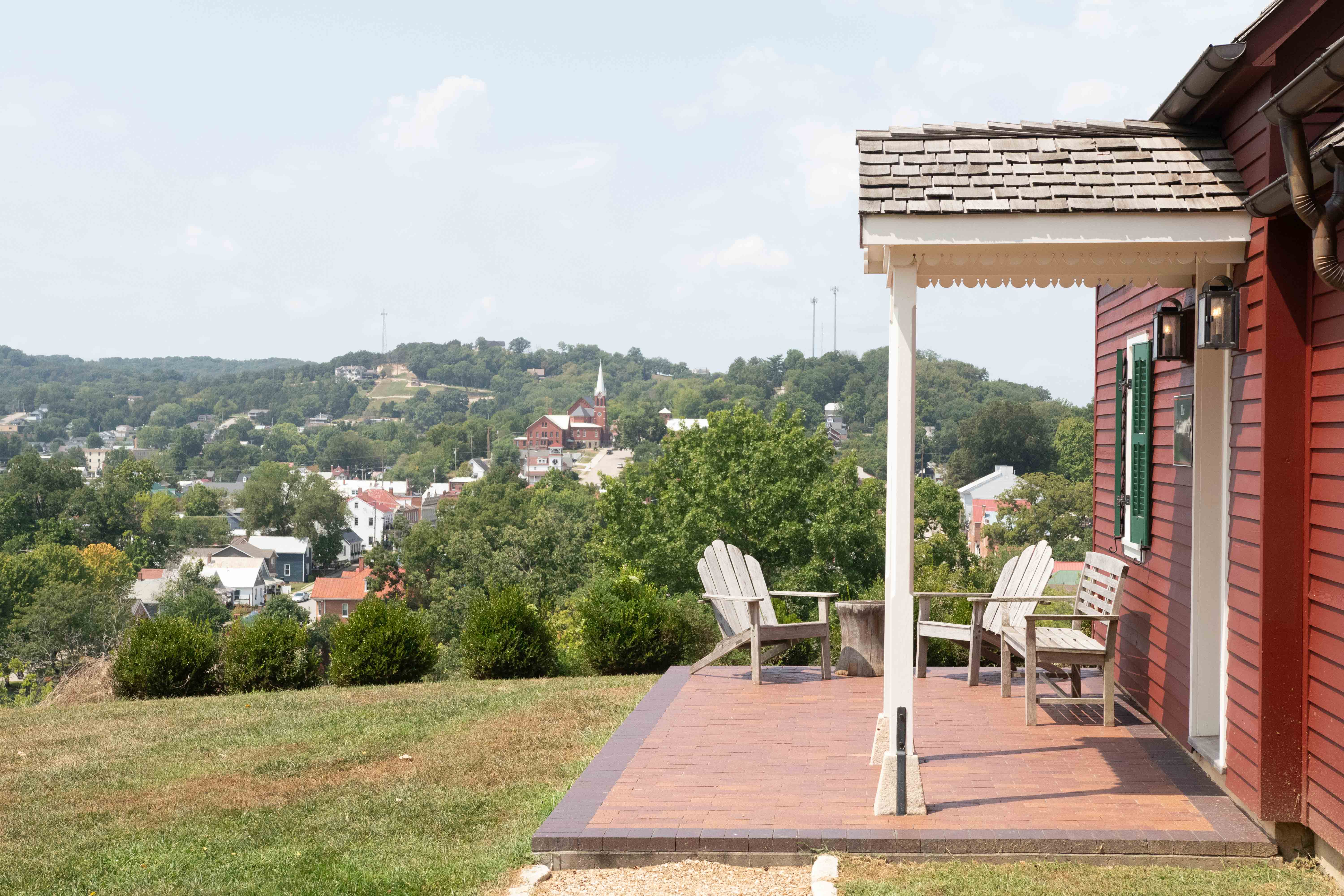 A red house with a porch overlooks a small town and green hills under a blue sky.