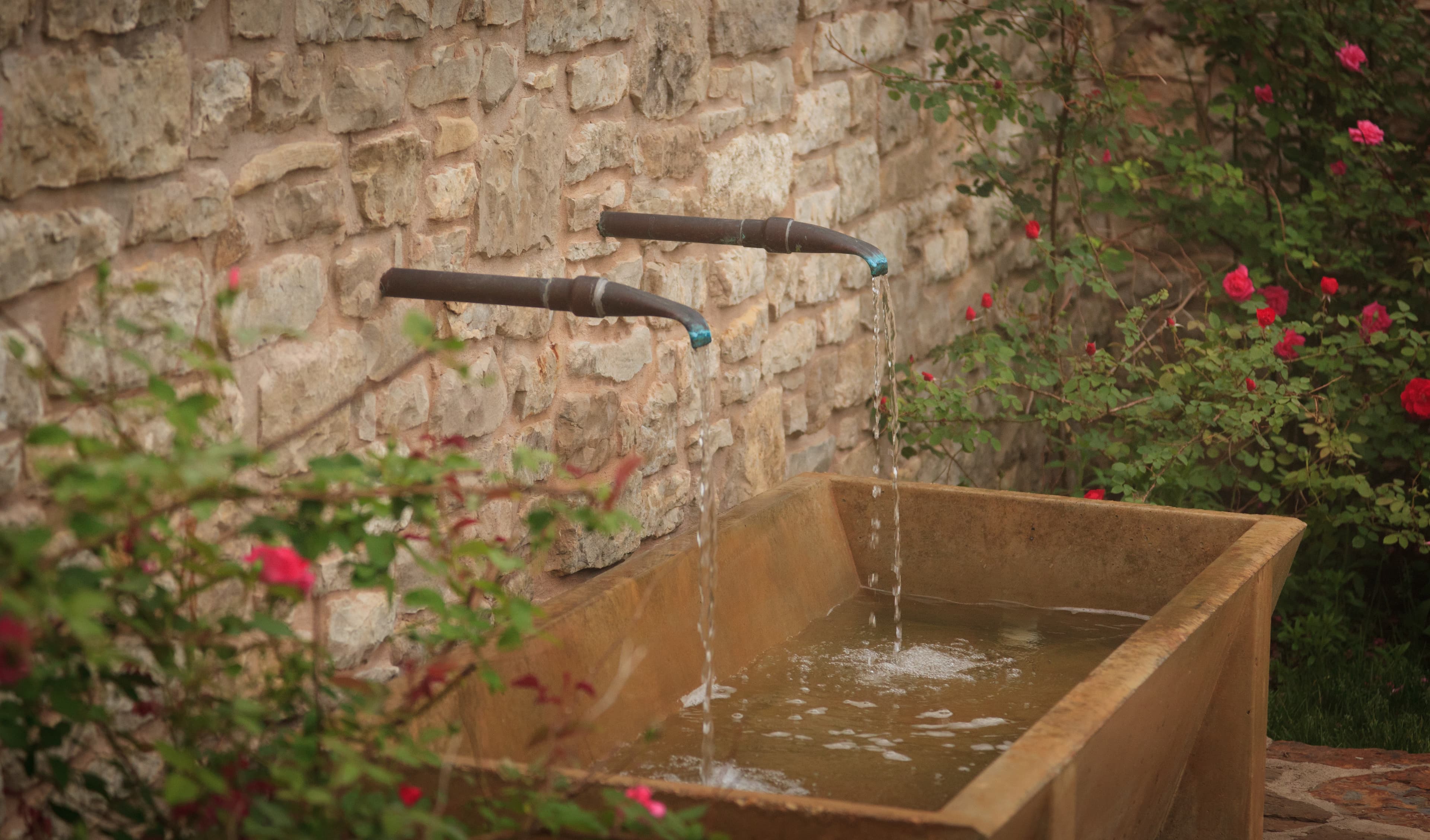 A rustic stone wall with two water spouts flowing into a stone basin surrounded by colorful flowers.