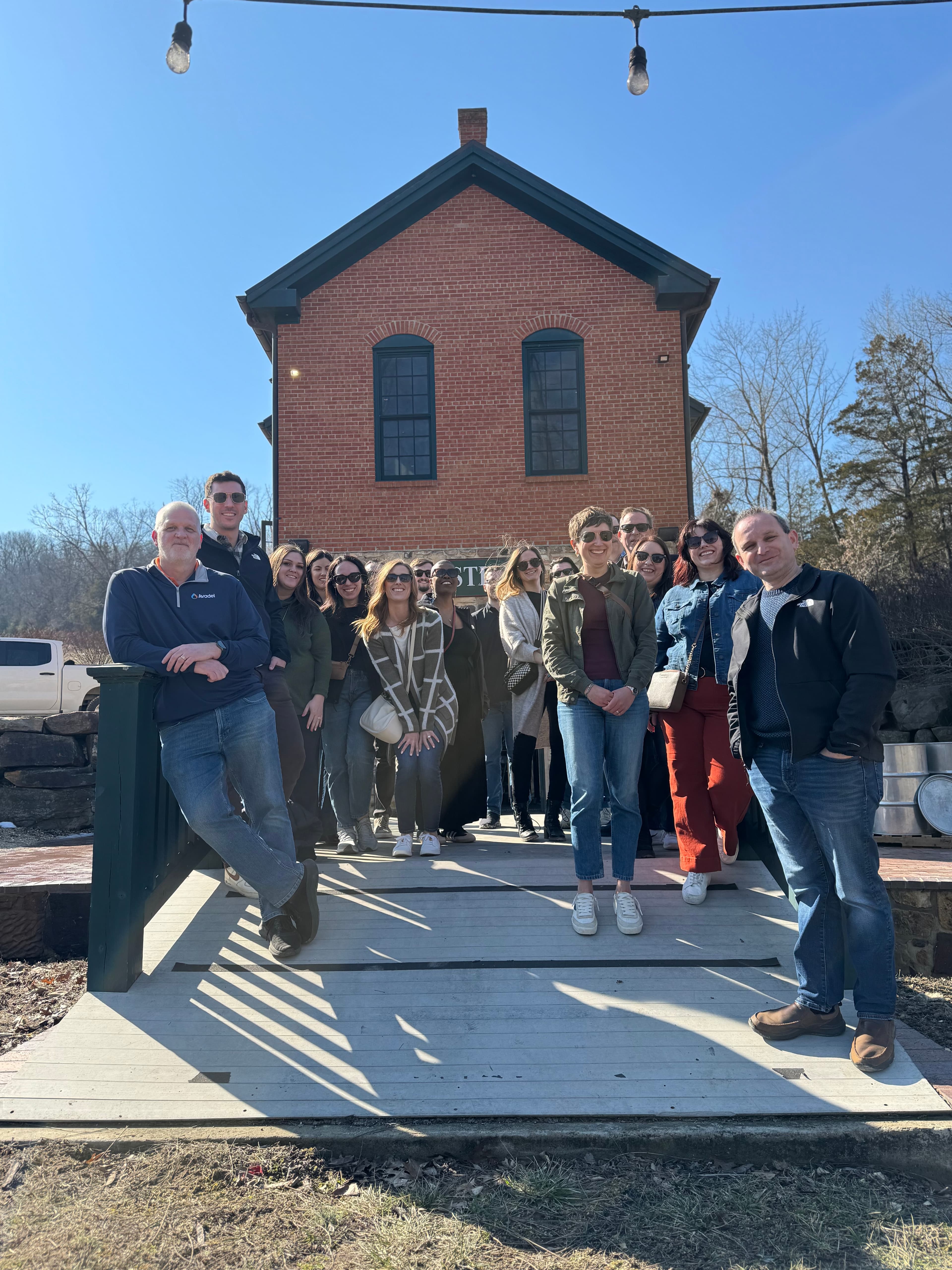 A group of people poses on a wooden platform in front of a brick building on a sunny day.