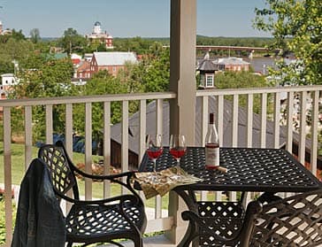 A cozy balcony table set with two glasses of red wine, a bottle, and snacks, overlooking a scenic view of a town and greenery.