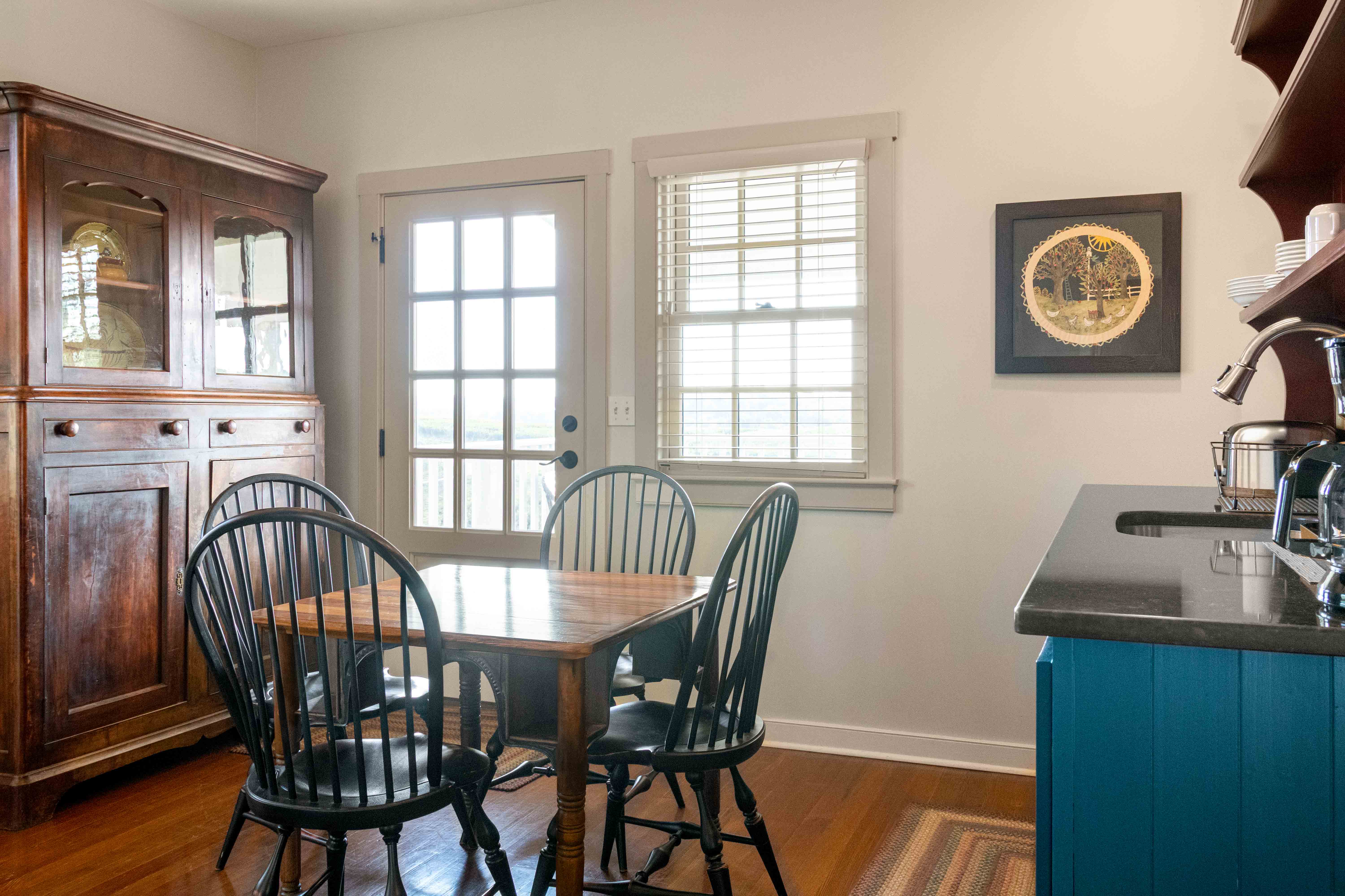 A cozy kitchen with a wooden dining table, four black chairs, an antique cabinet, and a blue kitchen counter.