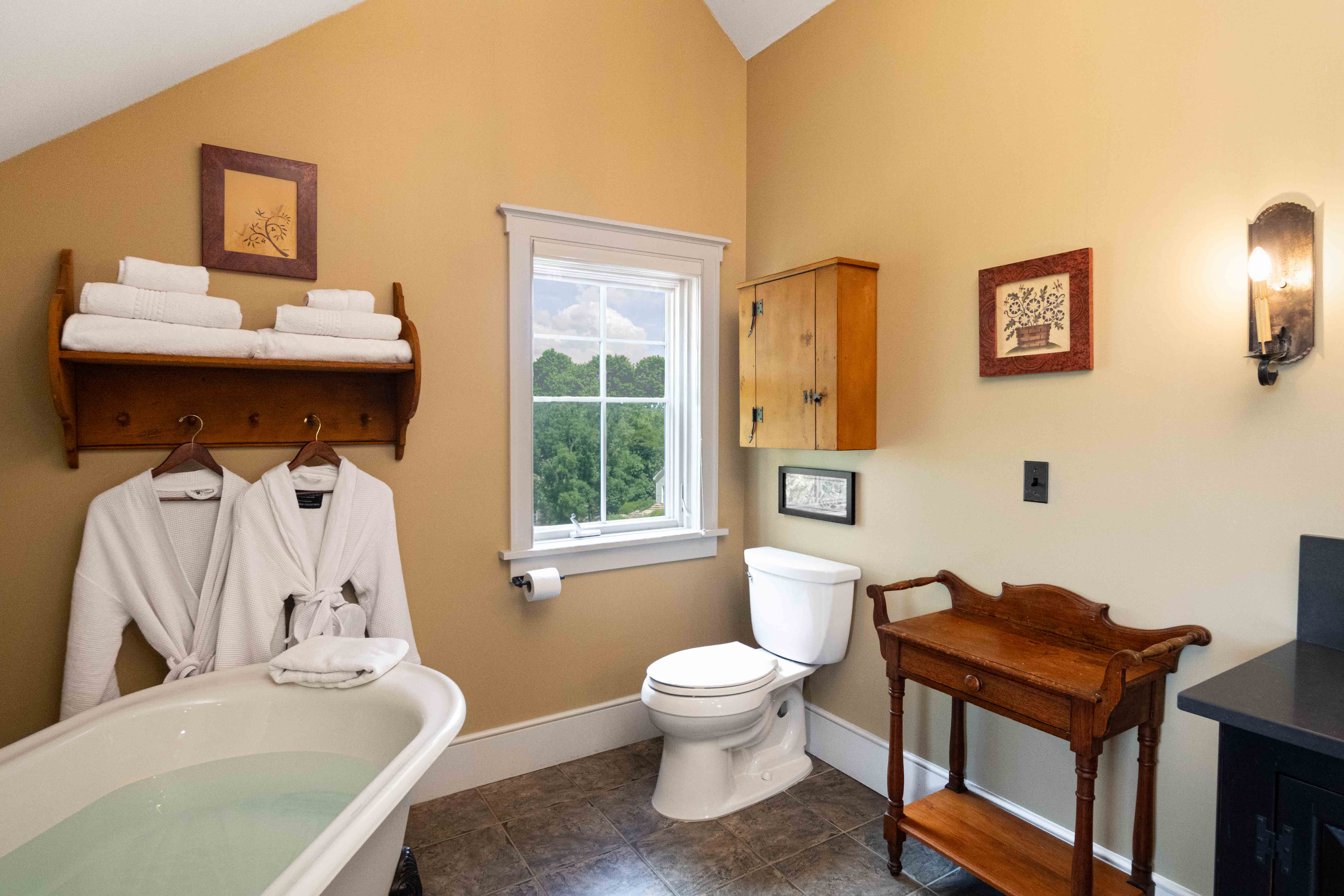A cozy bathroom featuring a freestanding tub, plush robes, and a window with a view of greenery.